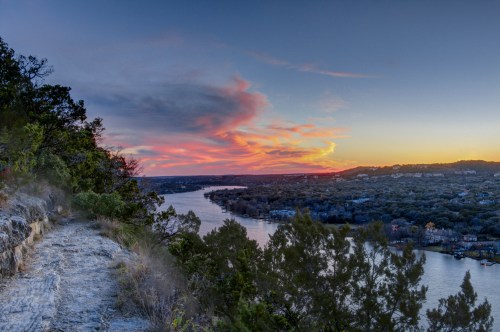 A scenic view of a river winding through a valley at sunset, with colorful clouds and lush greenery along the banks.