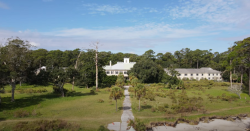 Aerial view of a white building surrounded by greenery and trees, with a path leading through the landscape.