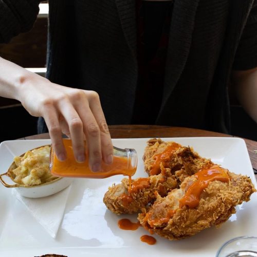 A hand drizzles hot sauce over two pieces of fried chicken, served with a side of macaroni and cheese.