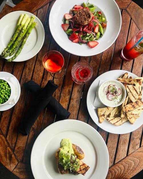 A wooden table with plates of salad, grilled fish, asparagus, peas, and dips, accompanied by colorful drinks.