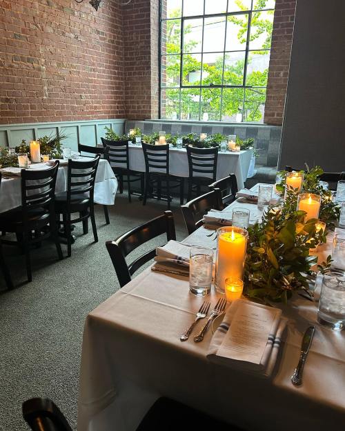 A cozy dining room with tables set for a meal, featuring candles and greenery, and large windows letting in natural light.