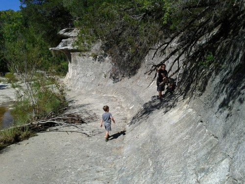 Two children play on a rocky path by a river, surrounded by trees and greenery. One child climbs while the other walks.
