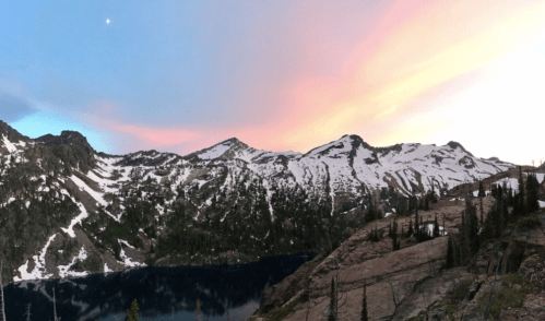 A panoramic view of snow-capped mountains under a colorful sunset sky, reflecting on a serene lake below.