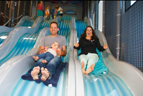A man and a woman slide down side-by-side water slides, laughing, with a child sitting on the man's lap.