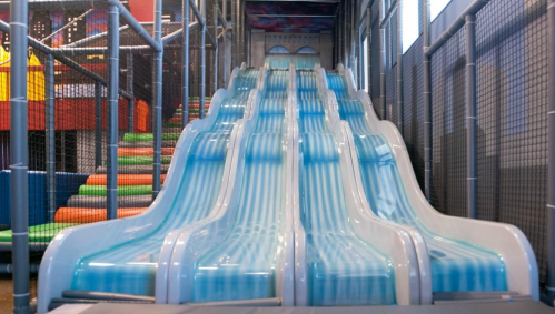 Two large, blue water slides in an indoor play area, surrounded by safety netting and colorful play structures.