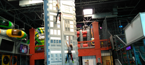 Two climbers ascend a tall, indoor climbing wall in a vibrant play area filled with colorful structures and slides.