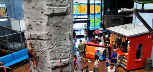 A busy indoor climbing gym with a rock wall, colorful seating, and a snack bar, filled with people enjoying activities.