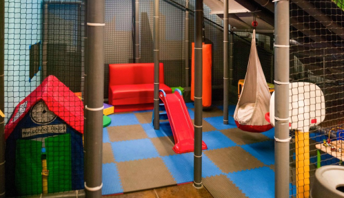 Indoor play area featuring a slide, swing, playhouse, and colorful mats, enclosed by safety netting.
