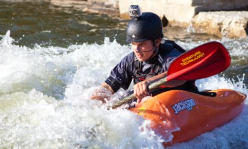 A kayaker navigates through whitewater rapids, wearing a helmet and a camera, with an orange kayak.