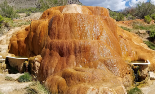 Colorful mineral deposits form a natural hot spring mound, surrounded by greenery and a blue sky.