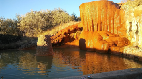 A natural hot spring with orange mineral deposits and a calm water surface, surrounded by dry vegetation and trees.