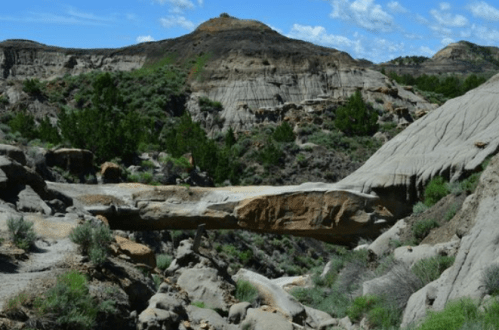 A rocky landscape with a natural stone bridge, surrounded by green shrubs and hills under a blue sky with clouds.