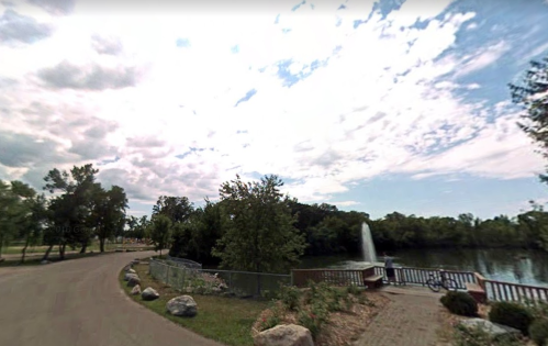 A scenic park view featuring a winding path, trees, and a fountain in a pond under a partly cloudy sky.