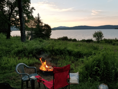 A cozy campfire surrounded by chairs, with a serene lake and mountains in the background at sunset.