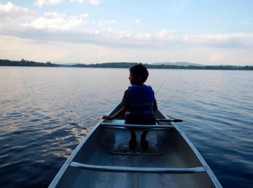 A child in a canoe faces a calm lake, wearing a life jacket, with trees and mountains in the background.