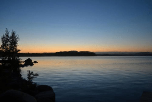 A serene lake at sunset, with calm waters reflecting the colorful sky and silhouettes of distant hills.