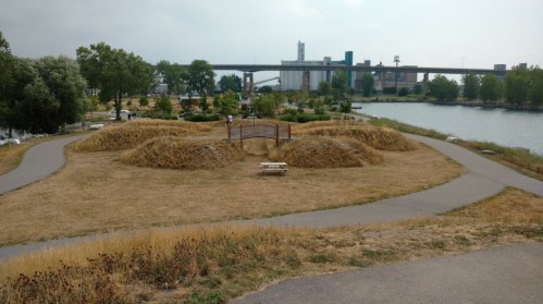 A park with dry grass, pathways, and a bench near a river, with industrial buildings in the background.