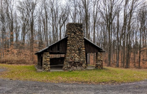 A rustic stone shelter with a chimney, surrounded by bare trees and a grassy area.