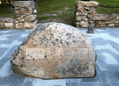 A large, weathered stone with the inscription "Miles Standish" on a paved surface, surrounded by stone structures.