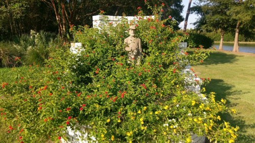 A statue partially hidden by vibrant green foliage and colorful flowers in a serene garden setting.