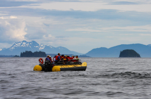 A group of people in a yellow inflatable boat on calm waters, with mountains and islands in the background.