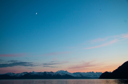 A serene twilight scene with a crescent moon over snow-capped mountains and a calm sea.