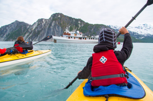 Two kayakers paddle in calm waters near a large boat and mountainous landscape, with snow-capped peaks in the background.