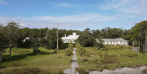 A scenic view of a white building surrounded by greenery and trees, with a pathway leading through the landscape.