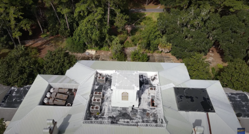 Aerial view of a building roof with air conditioning units and a white structure, surrounded by trees.