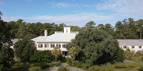 A large building surrounded by trees and greenery under a blue sky with scattered clouds.