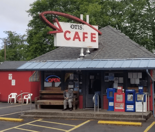 A small café with a retro sign reading "Otis' Café," featuring outdoor seating and an "Open" neon sign.