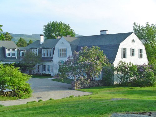 A charming white house with a sloped roof, surrounded by greenery and blooming flowers, set against a scenic backdrop.