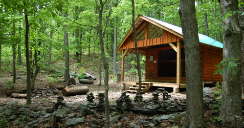 A wooden cabin surrounded by trees and rocks, with stacked stones in the foreground and a forested hillside in the background.