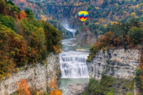 A colorful hot air balloon floats above a waterfall surrounded by vibrant autumn foliage and steep cliffs.