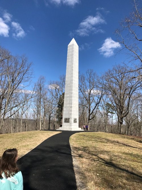 A tall white obelisk monument surrounded by trees and a clear blue sky, with a path leading up to it.