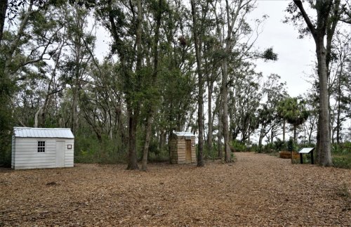 Two small wooden structures in a wooded area, surrounded by trees and a path leading into the distance.