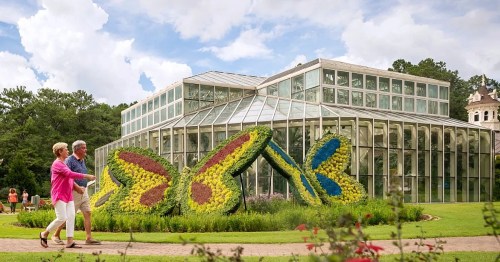 A couple strolls past a glass conservatory with colorful butterfly-shaped flower arrangements in a garden.