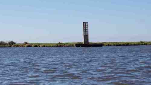 A tall structure stands on a platform in the water, surrounded by grassy land under a clear blue sky.