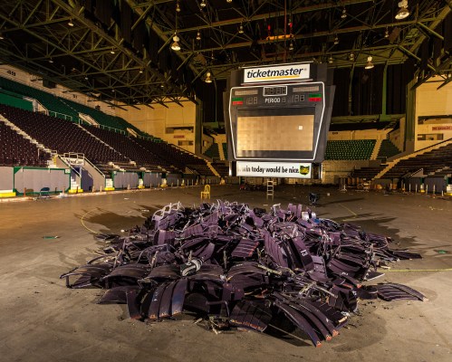 A pile of broken seats in an empty arena, with a large scoreboard displaying "Ticketmaster" in the background.