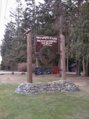 Sign for Sharpe Park in Montgomery-Dubany, surrounded by trees and grass, with decorative elements and stone base.