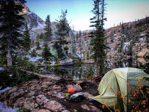 A tent set up by a serene lake, surrounded by tall trees and rocky terrain in a mountainous landscape.