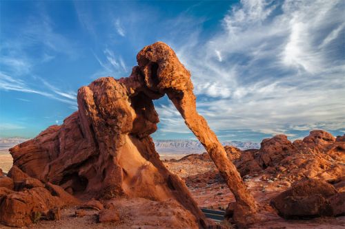 A striking rock formation resembling a figure, set against a blue sky with wispy clouds in a desert landscape.