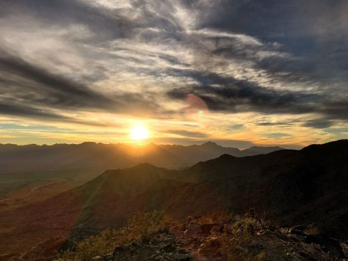 A stunning sunset over mountainous terrain, with vibrant clouds and a golden sun illuminating the landscape.