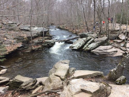 A serene river flows through a rocky landscape, surrounded by bare trees and a few distant hikers.