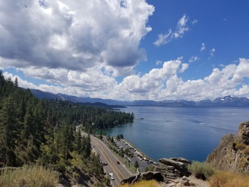 A scenic view of a lake surrounded by mountains and trees, with a road winding along the shoreline under a cloudy sky.