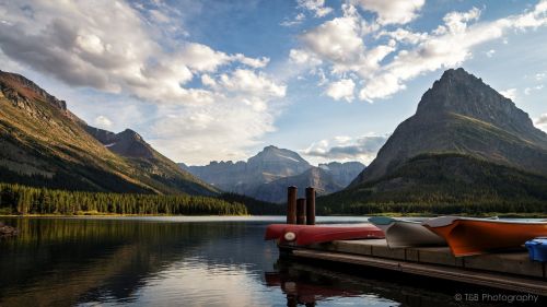 A serene lake surrounded by mountains, with colorful kayaks on a dock under a partly cloudy sky.