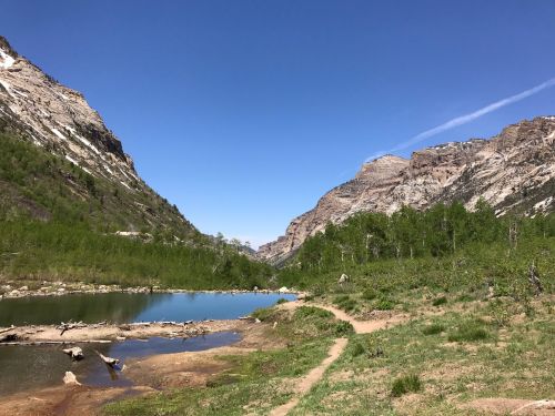 A serene landscape featuring a clear lake surrounded by green hills and towering mountains under a blue sky.