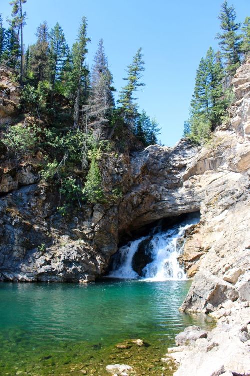 A serene waterfall cascades into a clear pool, surrounded by rocky cliffs and lush green trees under a blue sky.