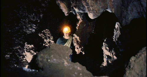 A person with a headlamp explores a dark cave, surrounded by rocky formations and shadows.