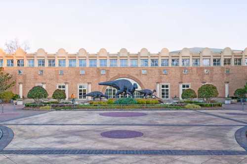 Exterior view of a brick building with a dinosaur sculpture in front, surrounded by landscaped gardens and pathways.